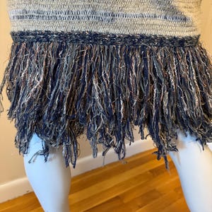 May include: A close-up of a woven top with a fringed hem. The top has horizontal stripes in grey, blue, and silver. The fringe is a mix of navy, silver, and copper threads, creating a textured and detailed appearance.