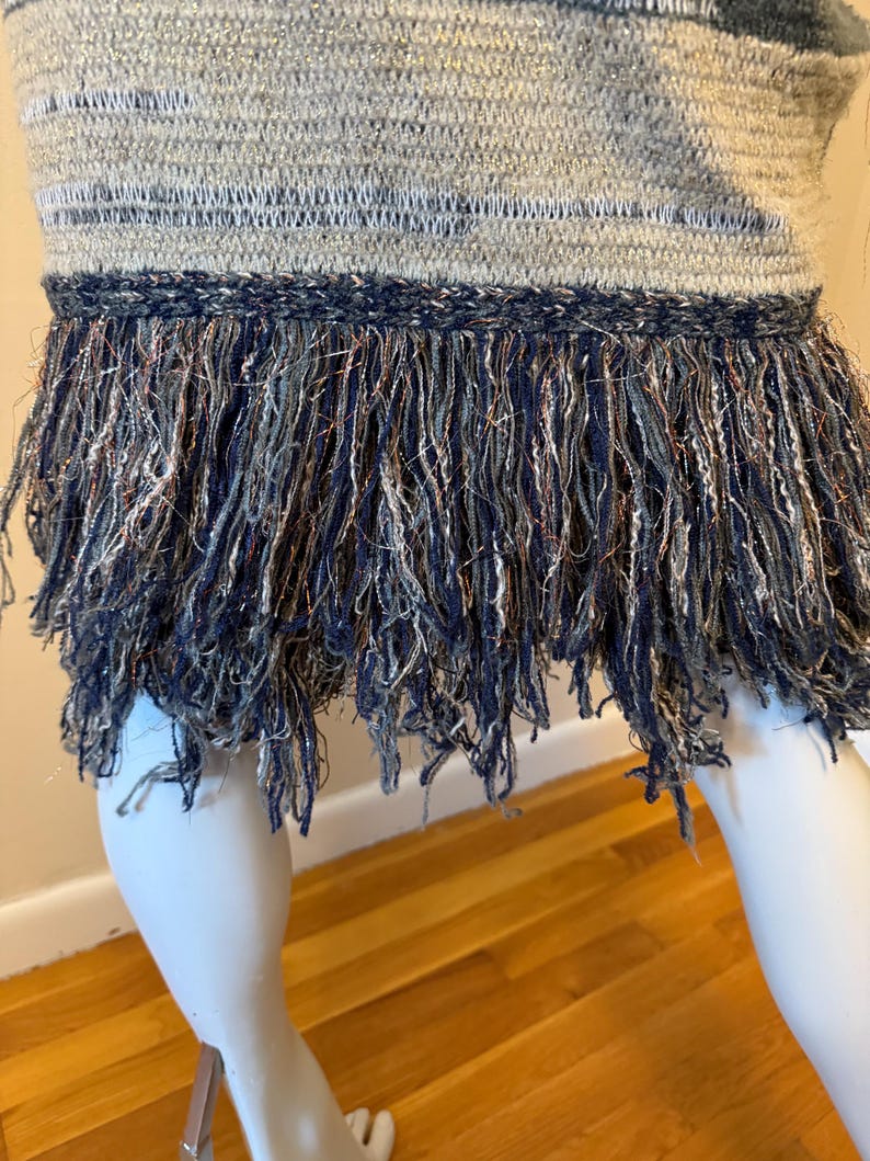May include: A close-up of a woven top with a fringed hem. The top has horizontal stripes in grey, blue, and silver. The fringe is a mix of navy, silver, and copper threads, creating a textured and detailed appearance.