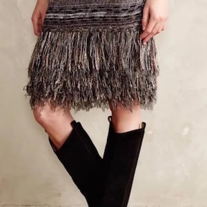 May include: A knitted mini-dress with a fringed hemline, featuring a gradient of colours from white to black. The dress is paired with black knee-high boots with a wooden heel. A gold bracelet is visible on the arm.