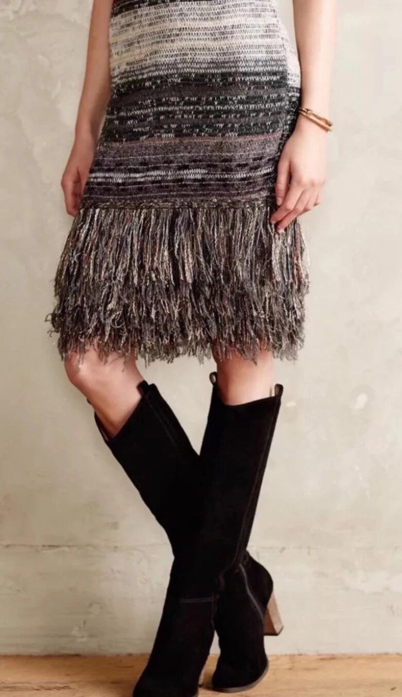 May include: A knitted mini-dress with a fringed hemline, featuring a gradient of colours from white to black. The dress is paired with black knee-high boots with a wooden heel. A gold bracelet is visible on the arm.
