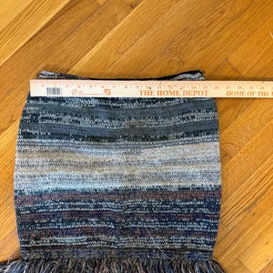 May include: A multi-coloured knitted skirt with a fringed hem. The skirt features horizontal stripes in shades of grey, blue, and brown. The skirt is approximately 40 cm wide.