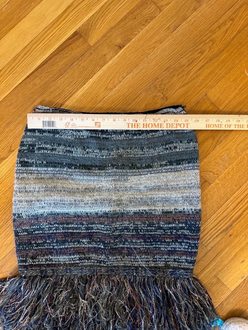 May include: A multi-coloured knitted skirt with a fringed hem. The skirt features horizontal stripes in shades of grey, blue, and brown. The skirt is approximately 40 cm wide.