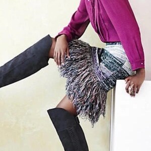 May include: A magenta long-sleeved shirt and a skirt with a fringe design. The skirt has a mix of colours, including white, green, and purple. The person is wearing black knee-high boots.
