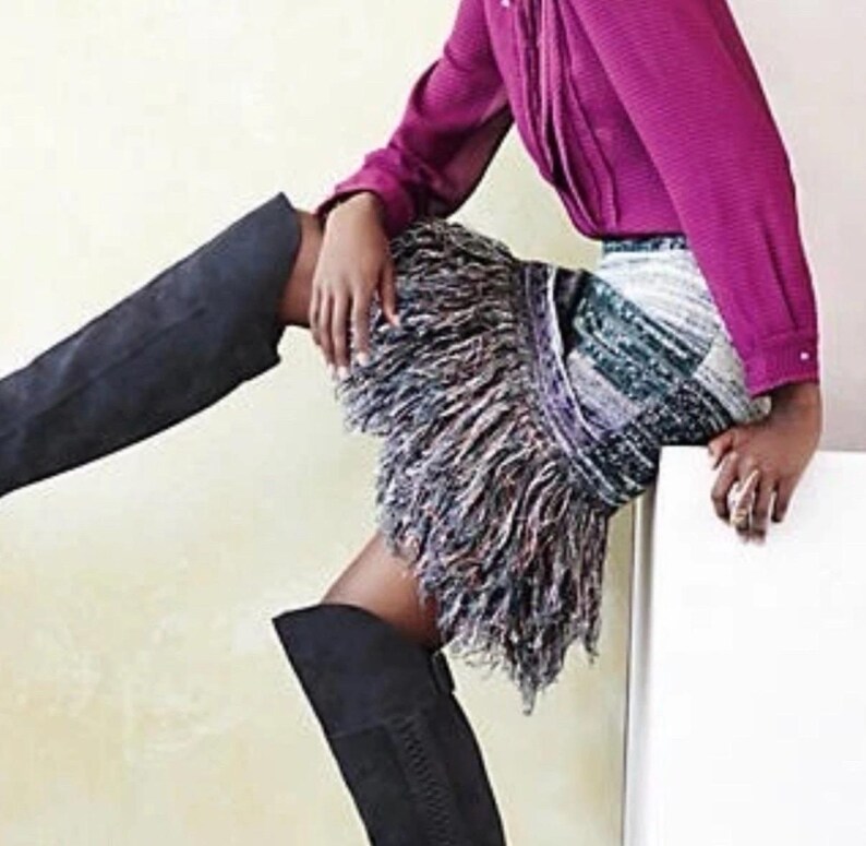 May include: A magenta long-sleeved shirt and a skirt with a fringe design. The skirt has a mix of colours, including white, green, and purple. The person is wearing black knee-high boots.