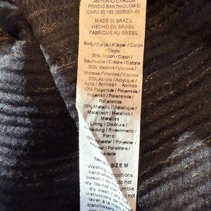 May include: A clothing label with text in multiple languages, detailing fabric composition and care instructions. The label includes percentages of cotton, viscose, acrylic, and metallic fibers. Washing instructions are also printed on the tag.