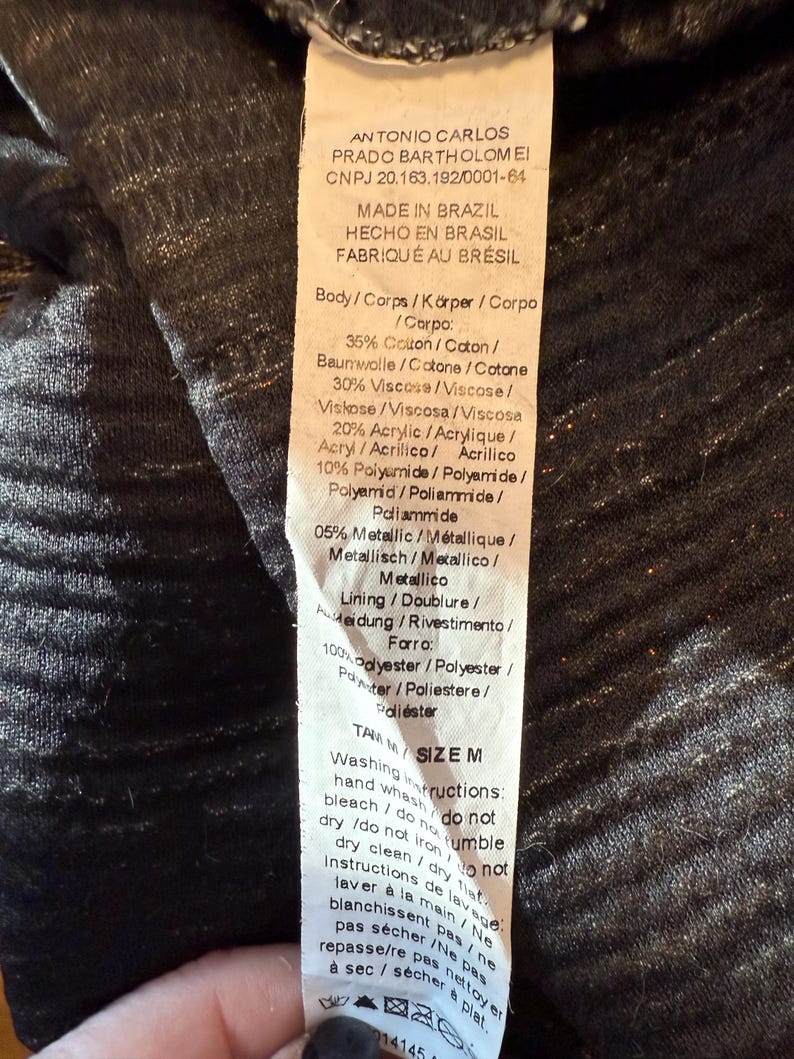 May include: A clothing label with text in multiple languages, detailing fabric composition and care instructions. The label includes percentages of cotton, viscose, acrylic, and metallic fibers. Washing instructions are also printed on the tag.