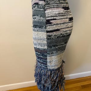 May include: A knitted pencil skirt with a patchwork design in shades of grey, white, and purple. The skirt features a fringe detail at the hemline with metallic threads. The skirt is displayed on a mannequin.