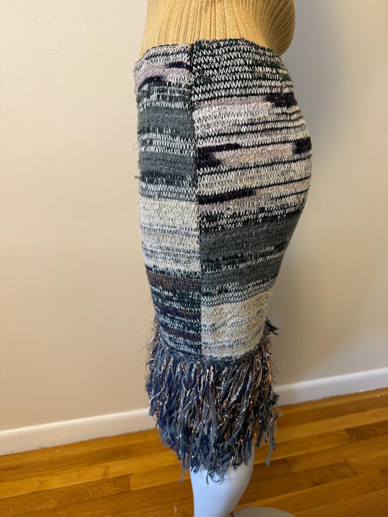 May include: A knitted pencil skirt with a patchwork design in shades of grey, white, and purple. The skirt features a fringe detail at the hemline with metallic threads. The skirt is displayed on a mannequin.