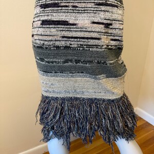 May include: A multi-coloured knitted skirt with a fringed hem. The skirt features a mix of grey, black, and beige horizontal stripes. The fringed hem is a dark blue with silver threads.