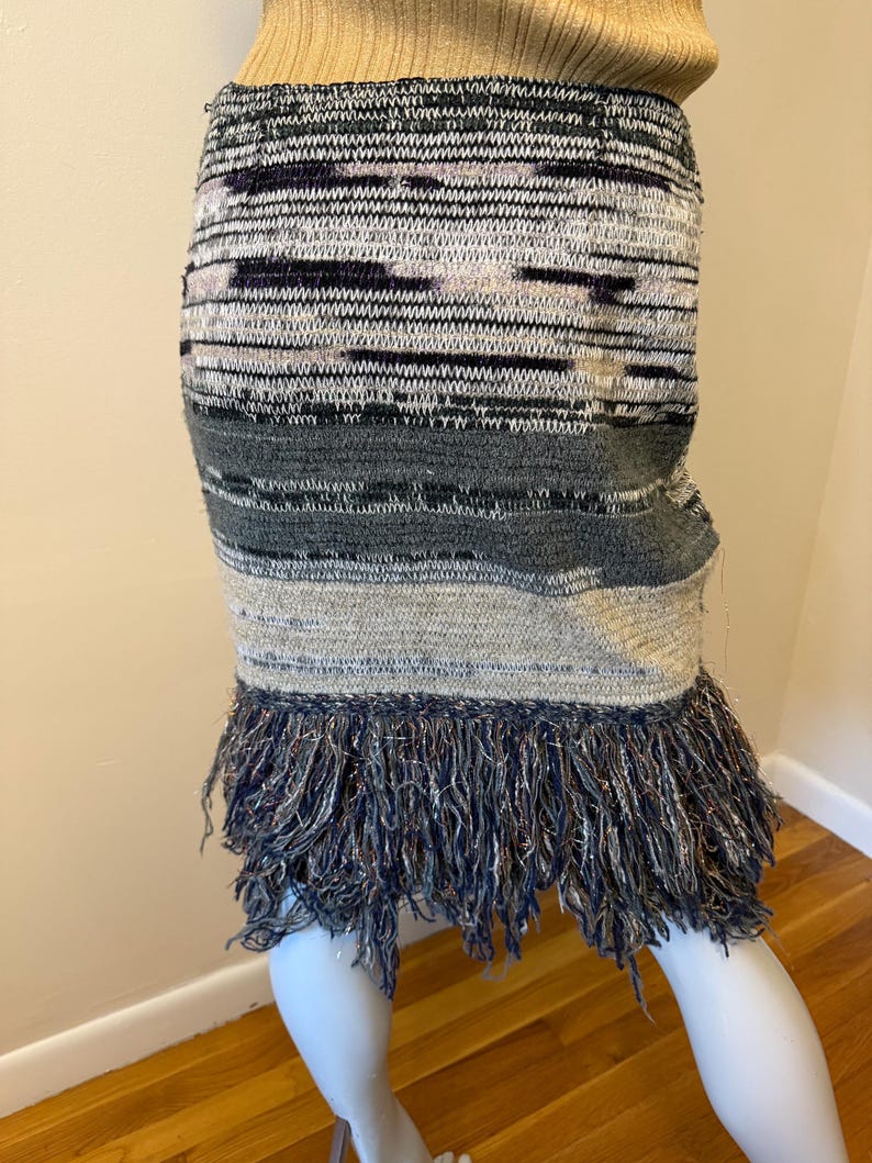May include: A multi-coloured knitted skirt with a fringed hem. The skirt features a mix of grey, black, and beige horizontal stripes. The fringed hem is a dark blue with silver threads.