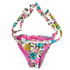 May include: Floral print bikini bottom with matching tie straps. The bikini features a vibrant pattern of pink, blue, and yellow roses on a white background. The edges are trimmed with white lace and a pink lining.
