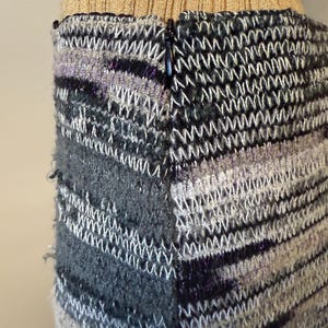 May include: Close-up of a knitted skirt with a zip. The skirt features a mix of grey, black, and white yarns, with a textured, woven pattern. The top portion of the skirt is a gold ribbed knit.