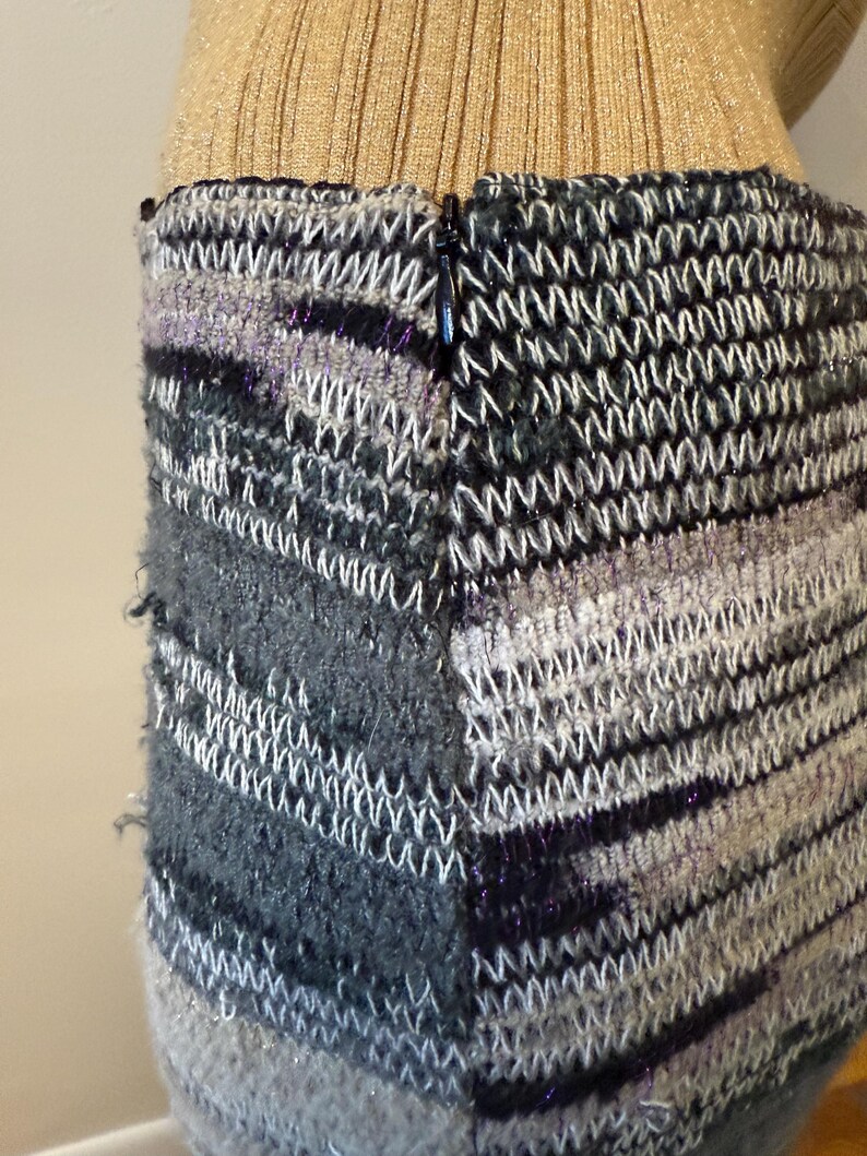 May include: Close-up of a knitted skirt with a zip. The skirt features a mix of grey, black, and white yarns, with a textured, woven pattern. The top portion of the skirt is a gold ribbed knit.