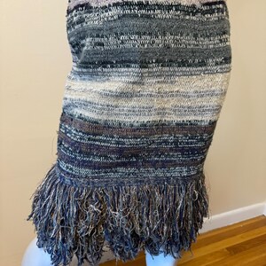 May include: A knitted skirt with a fringed hem. The skirt has horizontal stripes in shades of blue, grey, and white. The fringe at the bottom has a mix of blue and silver threads, adding texture and movement to the garment.