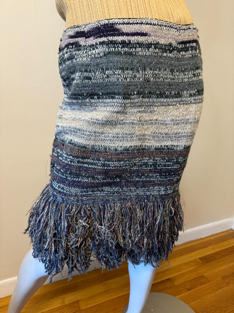 May include: A knitted skirt with a fringed hem. The skirt has horizontal stripes in shades of blue, grey, and white. The fringe at the bottom has a mix of blue and silver threads, adding texture and movement to the garment.