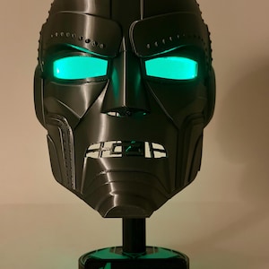 May include: A dark gray, 3D-printed mask with glowing green eyes, resembling a comic book character. The mask is mounted on a black stand with a base that reads "Avengers DOOMSDAY" in green and white lettering. The mask has a metallic appearance.