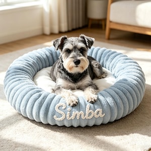 May include: A round, light blue pet bed with a ribbed, plush texture and a soft white center cushion. The front of the bed features the name Simba embroidered in white, textured script lettering. A small, grey and white dog is resting inside.