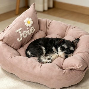 May include: A dusty pink pet bed with raised sides and a matching pillow featuring the embroidered white text "Joie" and a white daisy applique with a yellow centre. A small black and white dog is curled up sleeping inside the soft bed.