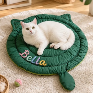 Puede incluir: Una alfombrilla acolchada para mascotas de color verde oscuro, con forma redonda, bordes superiores en forma de orejas de gato y una pequeña extensión similar a una cola. El nombre Bella está bordado con letras de lana coloridas.