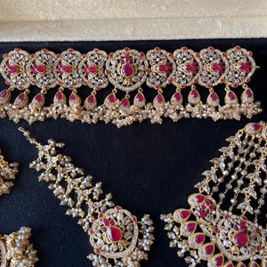 Op de afbeelding: Een goudkleurige sieradenset bestaande uit een brede choker-ketting, een paar grote oorbellen en een hoofdversiering. Elk stuk is versierd met druppelvormige rode stenen, heldere kristallen en kleine clusters van gebroken witte kralen.