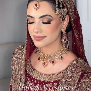 May include: A woman wearing a maroon and gold bridal ensemble. The outfit includes a headpiece, earrings, and a necklace with red gemstones. The dress is heavily embroidered with gold thread. The text "Hafsah Accessories" is visible on the dress.