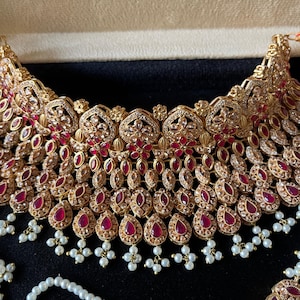 May include: A gold-toned choker necklace featuring intricate metalwork, embedded with numerous teardrop-shaped red stones and clear crystals. The lower edge is adorned with multiple dangling white pearl-like beads, creating a layered, ornate appearance.