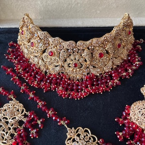 May include: A gold-toned choker necklace and matching earrings set featuring intricate metal filigree patterns, embedded with small clear crystals, and accented with deep red circular stones and dangling clusters of small red beads.