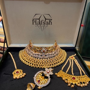 以下が含まれることがあります： Hafsah Accessoriesのロゴが入ったダークグリーンの箱に入ったゴールドトーンのジュエリーセット。クリアストーンと赤いアクセントが施された幅広で複雑なチョーカーネックレス、お揃いのイヤリング、マーンティッカ、マルチストランドのヘッドピースが含まれています。