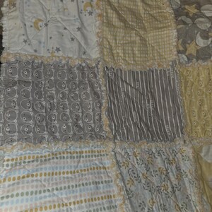 May include: A patchwork quilt with various patterned squares. The quilt features squares with moons and stars, bears, stripes, and floral designs in soft colors like yellow, gray, and white. The edges have a ruffled trim.