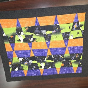 May include: A rectangular quilted table mat featuring a geometric pattern of orange, lime green, and purple fabric segments. The design includes Halloween-themed prints of ghosts, pumpkins, and stars, all framed by a solid black fabric border.