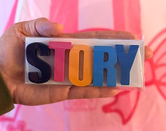 May include: A package containing the word "STORY" in colorful, raised letters. The letters are in navy blue, pink, orange, and blue. The background is a pink and white striped pattern.
