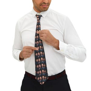 May include: A black necktie featuring a repeating pattern of a man's face. The tie is worn with a white dress shirt and black pants. The tie is being adjusted by the person wearing it.