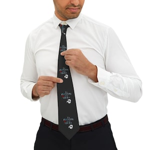 May include: Black tie featuring the text "The Phantom of the Opera" in white and a white mask design. The tie is being held by a person wearing a white dress shirt.