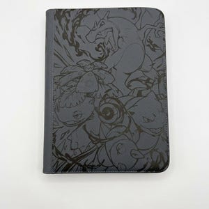 May include: A rectangular gray binder featuring a dark, monochromatic line art illustration of various Pokémon characters, including Charizard, Venusaur, and Blastoise, printed across the front cover with a textured, leather-like finish.