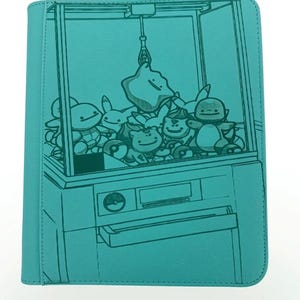 May include: Teal-colored notebook cover featuring a claw machine design. Inside the machine are various cartoon plush toys. The cover has a textured appearance and a simple, clean design, perfect for school or work.