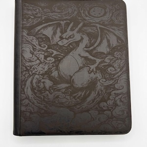May include: A black rectangular binder featuring a detailed, dark-toned embossed illustration of a dragon-like creature with wings, surrounded by swirling cloud patterns and a circular star-filled motif on the front cover surface.