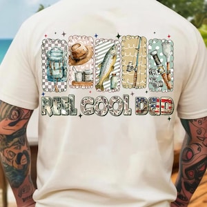 May include: Cream-colored t-shirt with a colorful graphic design featuring fishing-related images and the text "REEL COOL DAD." The design includes illustrations of fishing gear, a fish, and a hat.