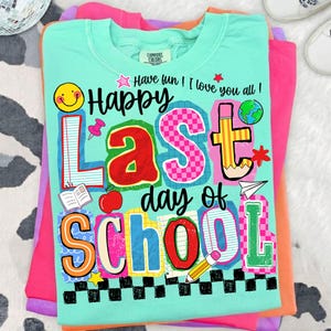 Pode incluir: Uma camiseta Comfort Colors verde claro com as palavras "Happy Last Day of School" em letras e gr&aacute;ficos coloridos. A camiseta tem um smiley, uma ma&ccedil;&atilde;, um l&aacute;pis e outras imagens com tema escolar. A camiseta est&aacute; dobrada em cima de outras camisetas coloridas.