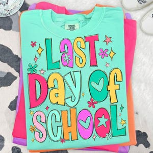 May include: A folded turquoise t-shirt with the words "LAST DAY OF SCHOOL" in colorful bubble letters. The shirt has a crew neck and is stacked on top of other folded shirts in pink, purple, and orange. Decorative stars and flowers surround the text.