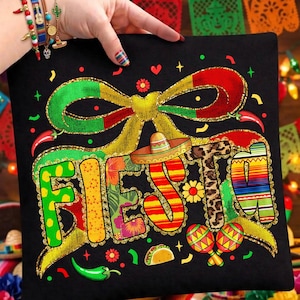 Puede incluir: Camiseta negra con la palabra "Fiesta" en letras coloridas y estampadas. El diseño incluye un sombrero, maracas, chiles y un lazo. La camiseta está sostenida por una persona con pulseras.