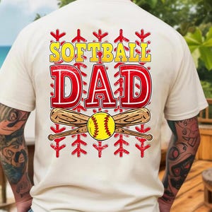 Op de afbeelding: Crèmekleurig T-shirt met de tekst "SOFTBALL DAD" in geel en rood. Het ontwerp bevat een softbal, twee gekruiste knuppels en rode stikseldetails, perfect voor een softbalfan.