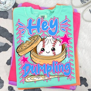 Puede incluir: Camiseta de color verde azulado claro con las palabras "Hey Dumpling" en escritura azul, con una bola de masa de dibujos animados con costuras de béisbol dentro de un vaporizador de bambú. Estrellas y acentos rosas y azules.