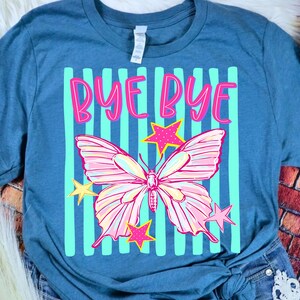 May include: Blue t-shirt with a graphic design featuring a pink and white butterfly, pink stars, and the words "BYE BYE" in pink. The design is set against a background of light blue vertical stripes.