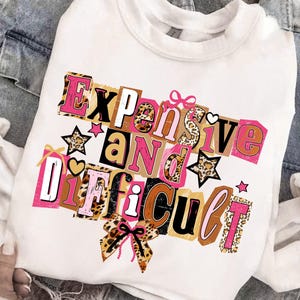 May include: White sweatshirt featuring the phrase "Expensive and Difficult" in a collage design. The text incorporates pink, brown, black, and leopard print patterns, with star and bow embellishments.