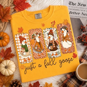 May include: A golden yellow t-shirt featuring a graphic of four geese dressed in autumn clothing, including hats and scarves, within rectangular frames. The text "just a fall goose" is printed in a cursive black font below the illustrations.