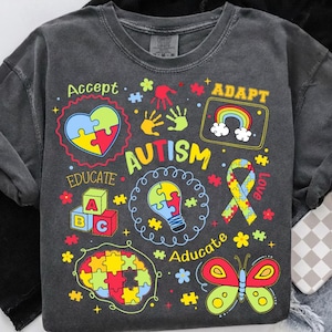 May include: Dark grey sweatshirt featuring autism awareness graphics. The design includes puzzle pieces, a rainbow, a ribbon, and the words "Accept," "Educate," "Adapt," and "Love."