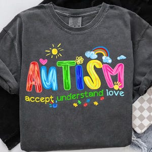 May include: Dark gray sweatshirt with the word "AUTISM" in colorful, bubble letters. The design includes a rainbow, sun, hearts, and the words "accept, understand love". A phone with a checkered case is visible.