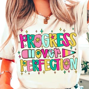 Può includere: T-shirt color crema con la scritta "PROGRESS OVER PERFECTION" in lettere colorate in stile retrò. Il design include stelle e fiori rosa, turchese, gialli e verdi. Una t-shirt grafica casual e d'ispirazione.