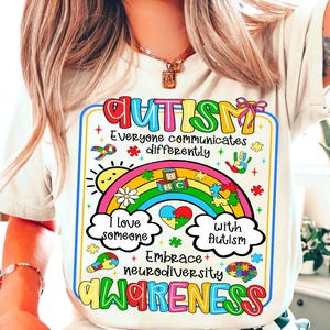 May include: White t-shirt with a colorful graphic promoting autism awareness. The design includes a rainbow, puzzle pieces, and the words "Autism Awareness." The shirt's message is "Everyone communicates differently."