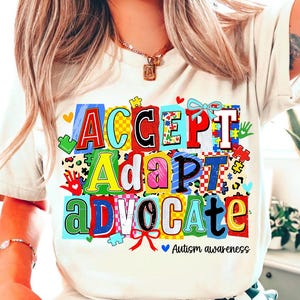 May include: Cream-colored t-shirt with the words "ACCEPT, ADAPT, ADVOCATE" in colorful block letters. The design includes puzzle pieces, hearts, and the text "Autism awareness".